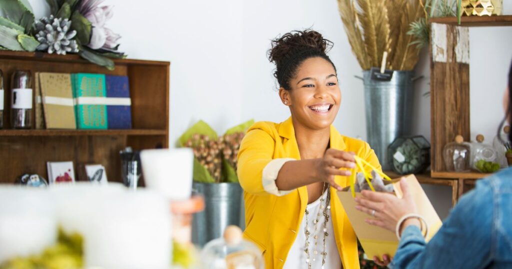 A woman in a yellow blazer smiles and she hands a customer a bag after checking out at a bookstore.