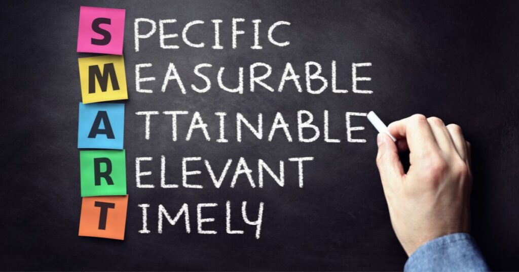 a person is writing on a chalkboard smart goals and explaining the acronym: Specific, measurable, attainable, relevant, timely.